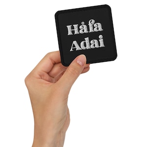 Peut inclure: Un patch carré noir avec du texte brodé blanc qui dit "Häfå Adai".