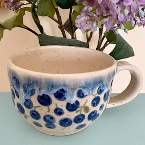 May include: A ceramic mug with a cream interior and a blue exterior. The mug has a blueberry design with blue and green accents. The handle is curved. The mug is on a light blue surface. Measurements are in cm.