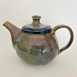May include: A ceramic teapot with a rounded body, a curved handle, and a spout. The glaze features a blend of deep blue, earthy brown, and moss green tones with a glossy finish. A matching lid with a small knob sits securely on the top opening.
