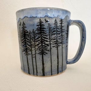 May include: A ceramic mug with a light blue and grey speckled glaze featuring a black ink illustration of a dense pine forest. The rim has a lighter blue drip effect, and the design includes small silhouettes of birds flying above the trees.