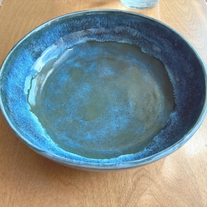 May include: A round ceramic bowl featuring a deep blue and black reactive glaze. The interior base has a smooth, muted olive-green finish that transitions into a vibrant, textured cobalt blue rim with darker charcoal-coloured edges and highlights.