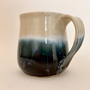 May include: A ceramic mug featuring a two-tone glaze design. The upper portion is a smooth, light beige, while the lower section transitions into a deep, dark blue with a white drip effect where the two colours meet. It has a curved handle.