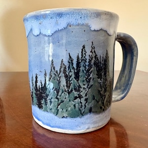 May include: A ceramic mug with a light blue glaze and a hand-painted forest scene featuring dark green pine trees. The mug has a sturdy handle and a smooth, cylindrical shape with a slightly flared rim, showcasing a speckled texture throughout the finish.
