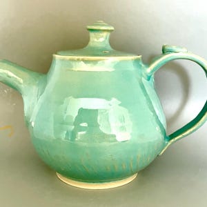 May include: A seafoam green ceramic teapot with a rounded body, curved handle, and spout. The lid has a small knob. The base and rim have a cream accent. A decorative item for the kitchen, this teapot is a functional piece.