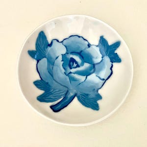 May include: A small, white ceramic dish featuring a hand-painted blue floral design. The flower is a peony with detailed petals and leaves. The dish is round with a slightly raised edge, ideal for small items.