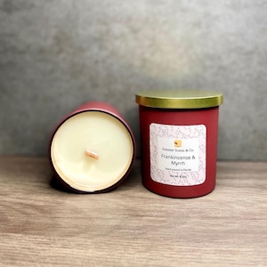 以下が含まれることがあります： 木製の表面に、金色の蓋が付いた2つの赤いキャンドルが置かれています。1つのキャンドルは横になっており、木の芯とクリーミーなワックスが見えます。もう1つのキャンドルには「Frankincense & Myrrh」と書かれたラベルが付いています。