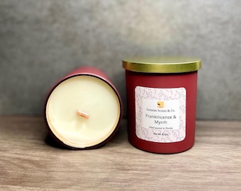 May include: Two red candles with gold lids are displayed on a wooden surface. One candle is lying on its side, revealing the wooden wick and creamy wax. The other candle has a label that reads "Frankincense & Myrrh".