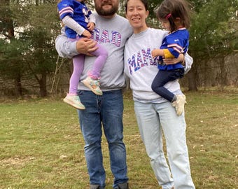 May include: A family of four poses outdoors. The parents wear gray sweatshirts with "DAD" and "MAMA" in purple letters. The children wear blue and white jerseys and are held by their parents. The family is standing on grass.