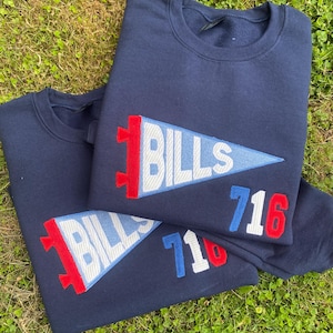 Puede incluir: Dos sudaderas azul marino con un diseño de banderín. El banderín es azul claro con la palabra "BILLS" en blanco y un borde rojo y blanco. El número "716" está bordado en rojo, blanco y azul.