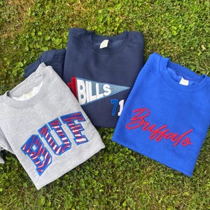 May include: Three crewneck sweatshirts featuring team-inspired designs. One is grey with the word BUT in blue and red striped lettering. One is navy blue with a pennant graphic reading BILLS and the number 71. One is royal blue with Buffalo in red.