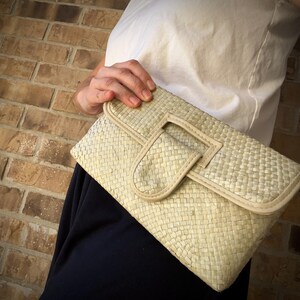 May include: A woven clutch bag with a flap closure and a white leather trim. The bag is made of a light beige material and has a woven texture.