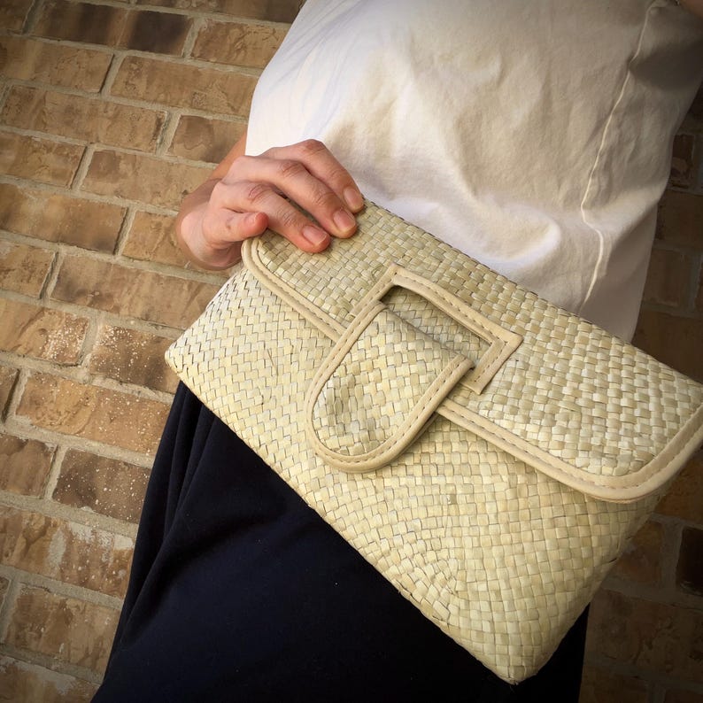 May include: A woven clutch bag with a flap closure and a white leather trim. The bag is made of a light beige material and has a woven texture.