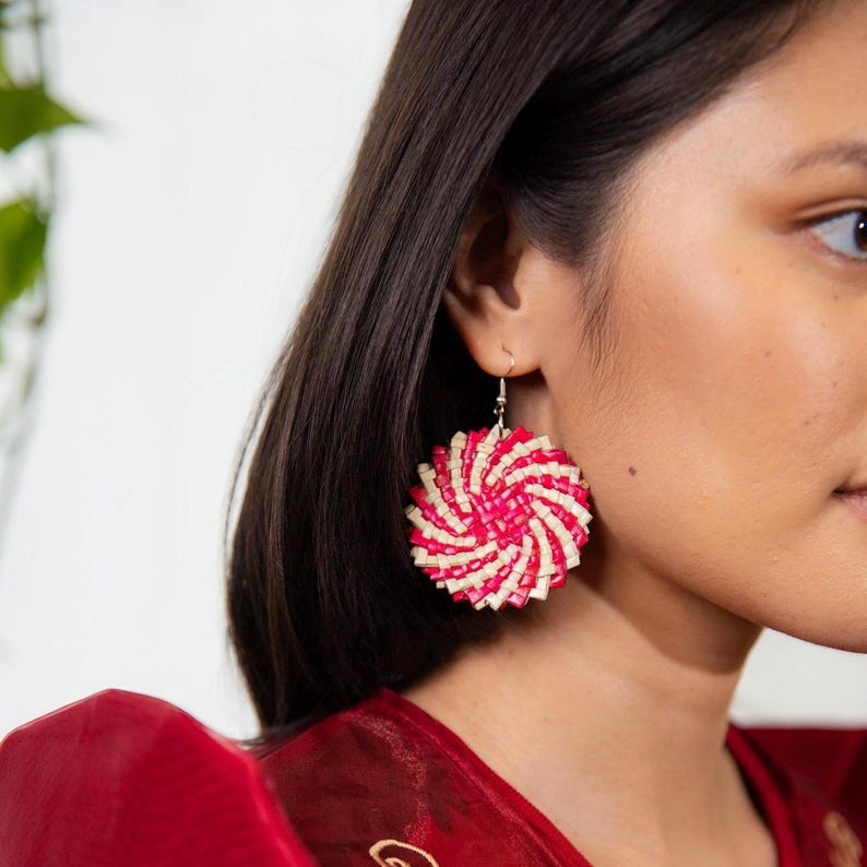 May include: A circular dangle earring featuring a woven spiral pattern in alternating bright pink and cream-colored strips. The earring is suspended from a simple silver-toned metal hook, creating a textured, round design that hangs from the earlobe.