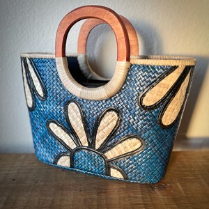 May include: A blue woven tote bag with a floral design. The bag features a wooden handle and a woven pattern in blue, cream, and black. The bag is approximately 12 inches wide and 10 inches tall.