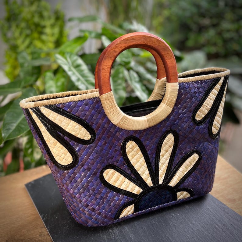May include: A woven tote bag with a purple and beige floral design. The bag features a wooden handle and a black zipper. The bag is made of woven material and has a rectangular shape.