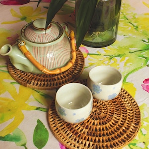 May include: A green and white striped teapot with a bamboo handle sits on a round woven rattan coaster. Two white teacups with blue floral designs are on similar coasters.