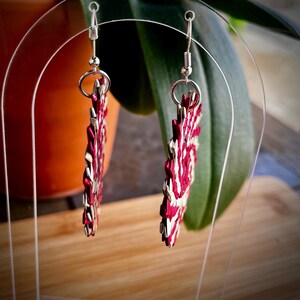 May include: A pair of dangle earrings featuring circular, woven components in alternating shades of deep magenta and metallic silver. The earrings are suspended from silver-toned metal fishhook wires, showcasing a textured, intricate pattern.
