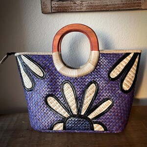 May include: A woven tote bag in shades of purple, featuring a floral design with black and cream accents. The bag has a wooden handle and a woven straw handle. The bag is designed with a zipper closure.