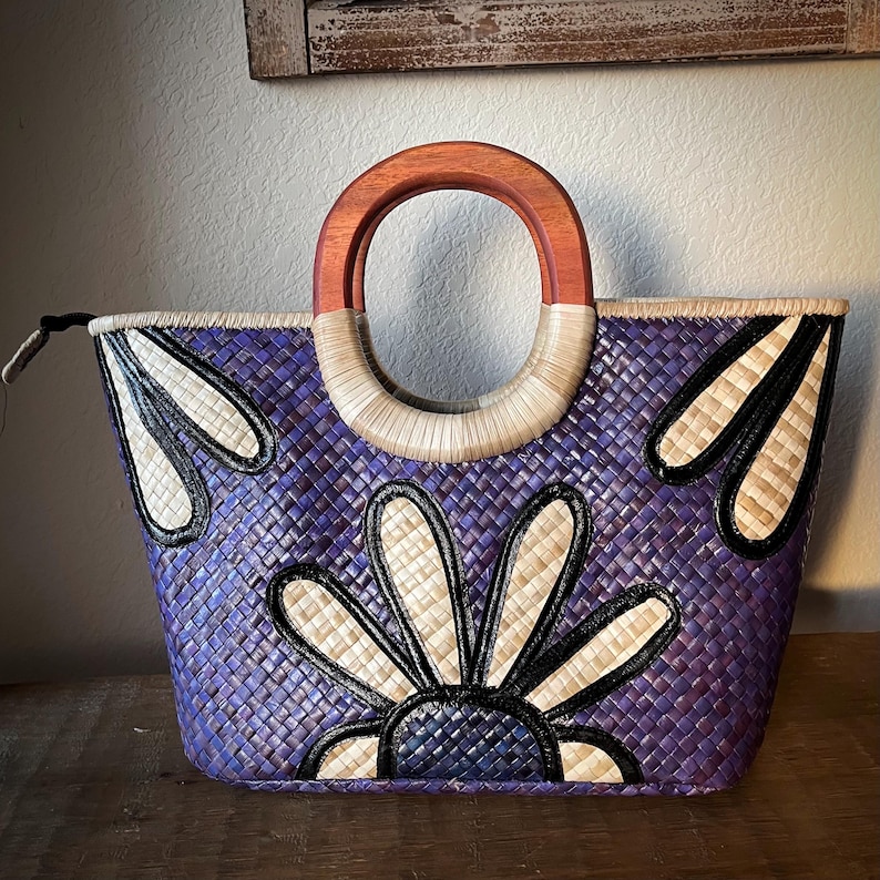 May include: A woven tote bag in shades of purple, featuring a floral design with black and cream accents. The bag has a wooden handle and a woven straw handle. The bag is designed with a zipper closure.