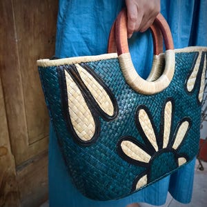 May include: A teal woven tote bag with a floral design. The bag features a woven pattern with light tan and black accents. It has wooden handles and a woven top edge. The bag is held by a person wearing a blue dress.