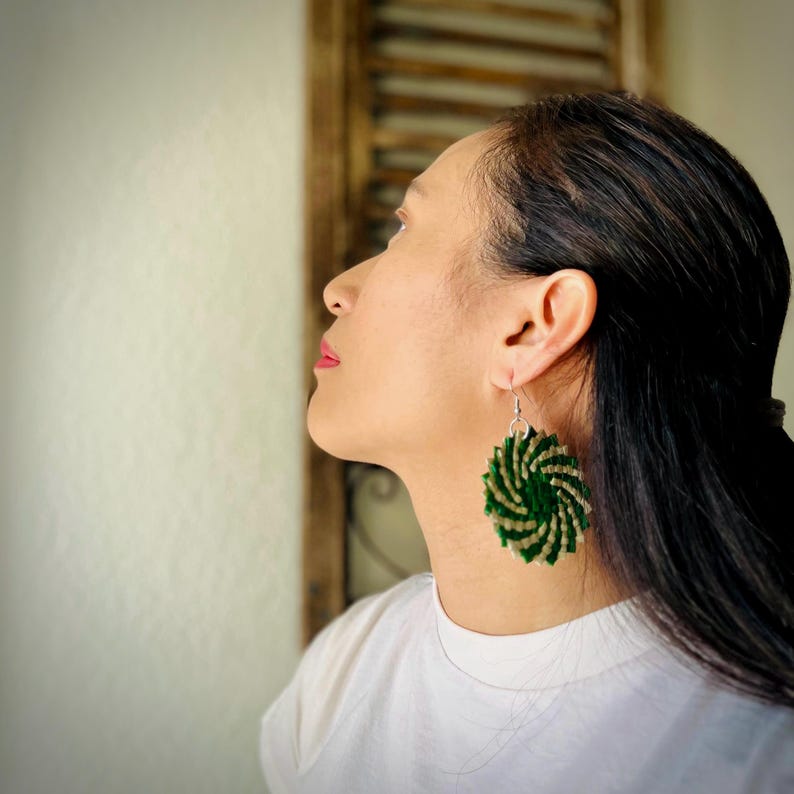 May include: A circular dangle earring featuring a spiraling pattern of alternating dark green and cream-colored woven fibers. The earring is attached to a silver-toned metal hook and hangs from the earlobe, displaying a textured, layered design.