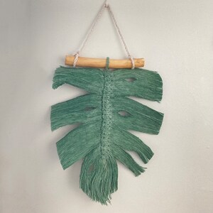 May include: A green macrame leaf wall hanging with a wooden dowel at the top. The leaf is made of yarn and has a fringed edge.
