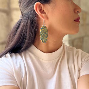 May include: An oval-shaped dangle earring featuring a textured, woven pattern in alternating shades of mustard yellow and slate blue. The earring is suspended from a simple silver-toned metal hook, creating a distinct, layered appearance.