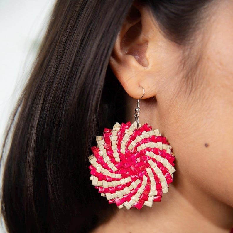 May include: A circular dangle earring featuring a woven pattern of vibrant hot pink and natural beige strips. The earring is suspended from a silver-toned metal hook and displays a radiating spiral design that creates a textured, layered appearance.