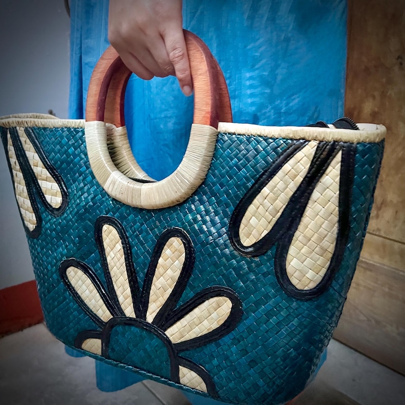 May include: A teal woven tote bag with a floral design. The bag features a woven pattern, wooden handles, and decorative accents in black and cream. The handles are made of wood and are a reddish-brown color. The bag is held by a person.