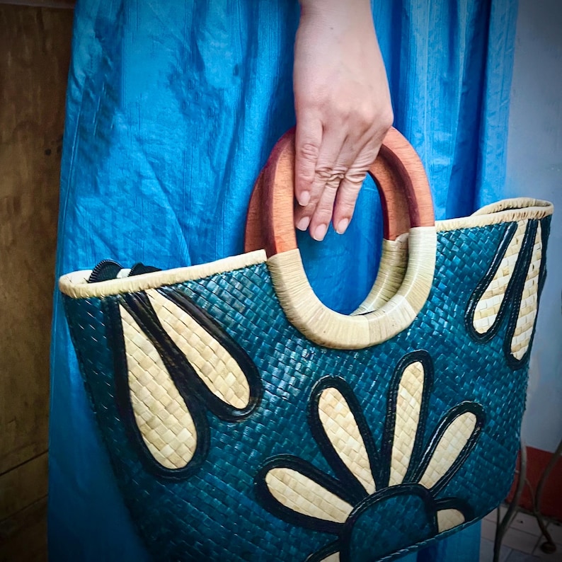 May include: A woven tote bag in teal with a floral design in black and natural tones. The bag features wooden handles and a woven rim. The bag is held by a person wearing a blue dress.