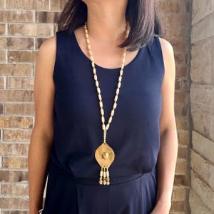 May include: A long, cream-colored beaded necklace with a gold pendant. The pendant is a large, flat, teardrop shape with a smaller circle in the center. The necklace is worn by a person with dark hair.