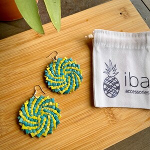 May include: A pair of round, woven earrings featuring a spiral pattern of teal and yellow strips, attached to silver-toned metal hooks. Beside the earrings is a small white fabric drawstring pouch with a black pineapple icon and the text iba accessories.