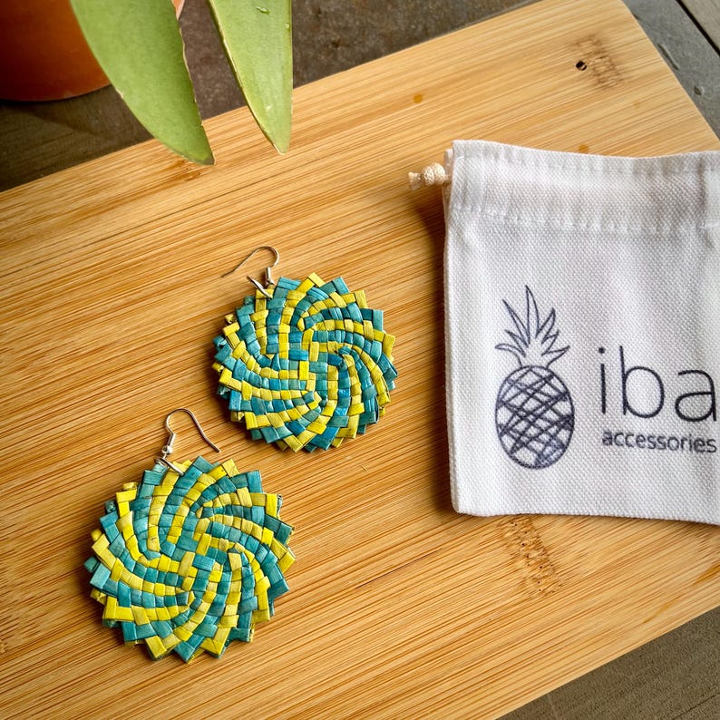 May include: A pair of round, woven earrings featuring a spiral pattern of teal and yellow strips, attached to silver-toned metal hooks. Beside the earrings is a small white fabric drawstring pouch with a black pineapple icon and the text iba accessories.