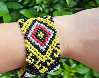 May include: A wide beaded cuff bracelet featuring a geometric pattern of small yellow, black, white, and red glass beads. The design consists of a central diamond motif surrounded by repeating black and yellow triangular shapes in a woven structure.