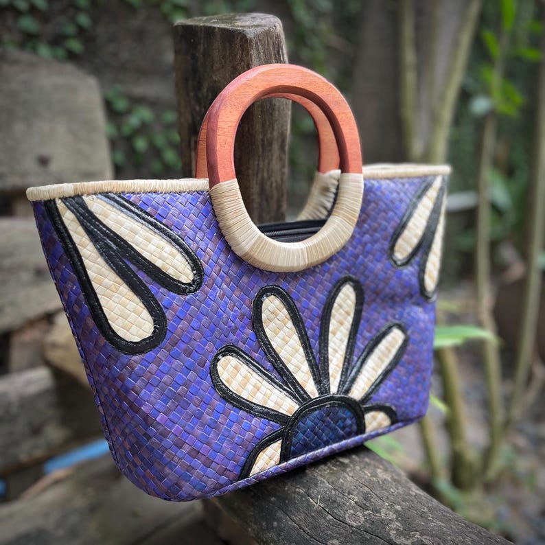 May include: A woven tote bag with a purple and blue floral design. The bag features a wooden handle and a woven straw trim. The floral design is in white and black. The bag is sitting on a wooden surface.