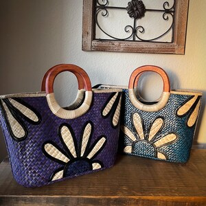 May include: Two woven tote bags with wooden handles. One bag is a deep purple with a white and black floral design, and the other is teal with a similar design. The handles are a light brown color.