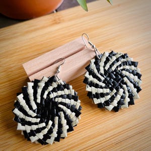 May include: A pair of circular earrings featuring a woven pattern of black and off-white strips arranged in a radiating spiral design. Each earring is attached to a silver-toned metal hook for hanging, creating a distinct geometric appearance.
