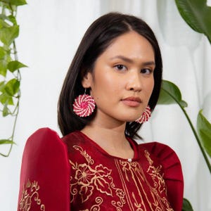 May include: Round earrings featuring a spiral pattern of small red and white beads. The earrings are worn with a deep red garment that has structured, pleated shoulders and intricate gold floral embroidery across the front chest area.
