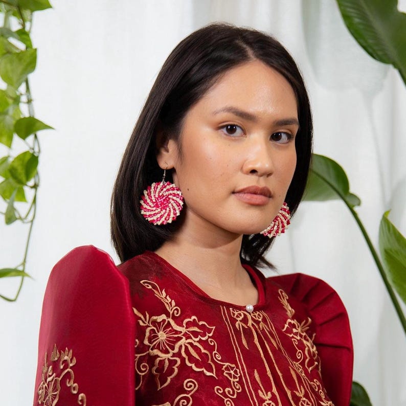 May include: Round earrings featuring a spiral pattern of small red and white beads. The earrings are worn with a deep red garment that has structured, pleated shoulders and intricate gold floral embroidery across the front chest area.