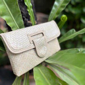 May include: A woven clutch bag with a light beige color and a flap closure. The bag is made of natural materials and has a subtle texture.