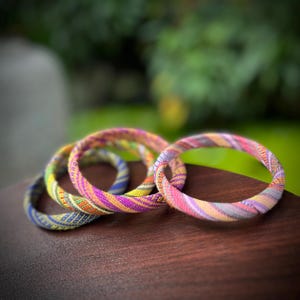 May include: Four colorful fabric bracelets in shades of blue, yellow, pink, and purple are arranged on a dark wooden surface. The bracelets have a striped pattern and appear to be handmade. The background is blurred, with green foliage visible.