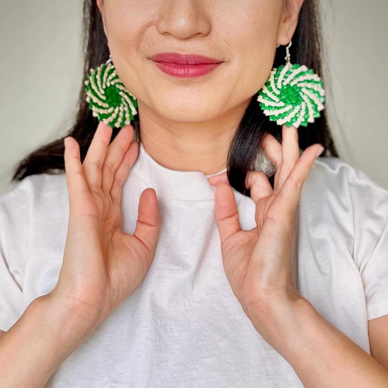 May include: A pair of circular earrings featuring a spiraling pattern of alternating bright green and off-white woven strips. The earrings have a round, disc-like shape with a textured, layered appearance and are attached to silver-toned hooks.