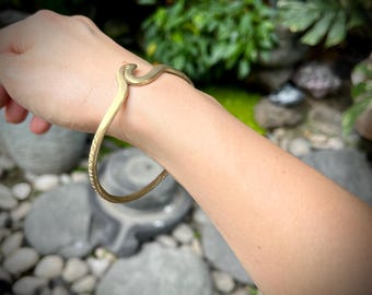 May include: A gold-colored snake-shaped bracelet. The bracelet curves around the wrist, with the snake's head at the top. The bracelet has a textured design along the bottom curve. The background is blurred, with greenery and rocks.
