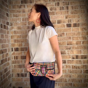 May include: A colorful woven clutch bag with a black buckle closure. The bag is held by a person wearing a white t-shirt and black pants.