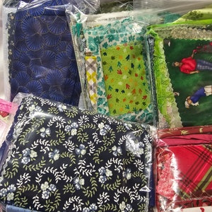 Op de afbeelding: Een verzameling stoffenresten in verschillende patronen en kleuren, waaronder blauw, groen, rood en roze. De stoffen zijn verpakt in doorzichtige plastic zakken.