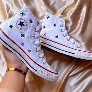 May include: White high-top trainers with green shamrock embroidery. The shoes feature a red and navy stripe on the sole and the classic Converse All Star logo. The trainers are displayed on a gold satin fabric.