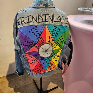 May include: A light-wash denim jacket with a colorful, geometric design on the back. The design features a central circle with radiating segments in various colors, including blue, purple, green, and red. The jacket is displayed on a black mannequin.