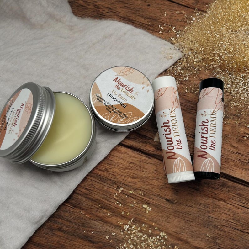 May include: A set of three lip balms in a tin and two tubes. The lip balms are labeled "Nourish the Dermis" and are unscented. The lip balms are on a wooden surface with a white cloth and a pile of brown sugar.