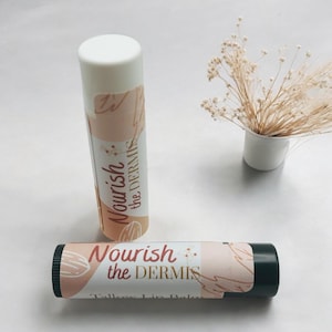 May include: Two lip balm tubes with a white and black base and a label that says "Nourish the Dermis. Tallow Lip Balm". The label has a peach and brown background with a floral design.