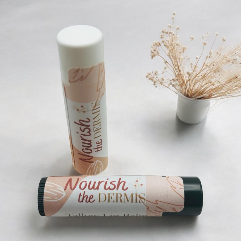 May include: Two lip balm tubes with a white and black base and a label that says "Nourish the Dermis. Tallow Lip Balm". The label has a peach and brown background with a floral design.